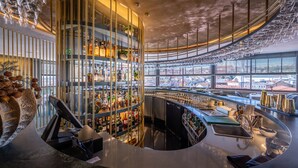 Un bar moderno con una barra curvada, varias estanterías llenas de botellas y vistas de la ciudad a través de amplios ventanales.