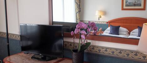 Habitación de hotel con una televisión sobre un escritorio de madera, una orquídea en maceta, un cuadro en la pared y una cama con sábanas blancas y almohadas azules.