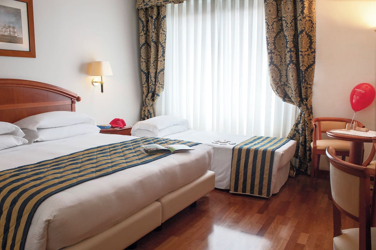 A hotel room with two beds, a bedside table with a lamp, a chair, a small table with a balloon, and a window with curtains.