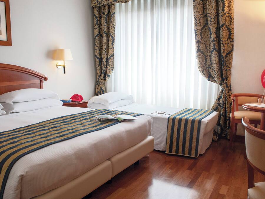 A hotel room with two beds, a bedside table with a lamp, a chair, a small table with a balloon, and a window with curtains.