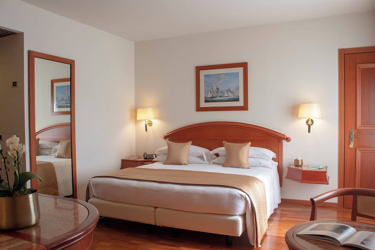 A hotel room with a large bed, bedside lamps, a bedside table, a chair, and a small table with a book.