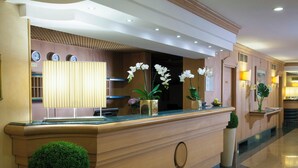 A hotel reception area with a wooden counter, a reception desk, and a wall-mounted clock. There are potted plants and a small display of fruits on the counter. - Starhotels Vespucci (Campi Bisenzio)
