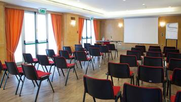 A conference room with rows of black chairs with red cushions, a whiteboard, and a projector screen.