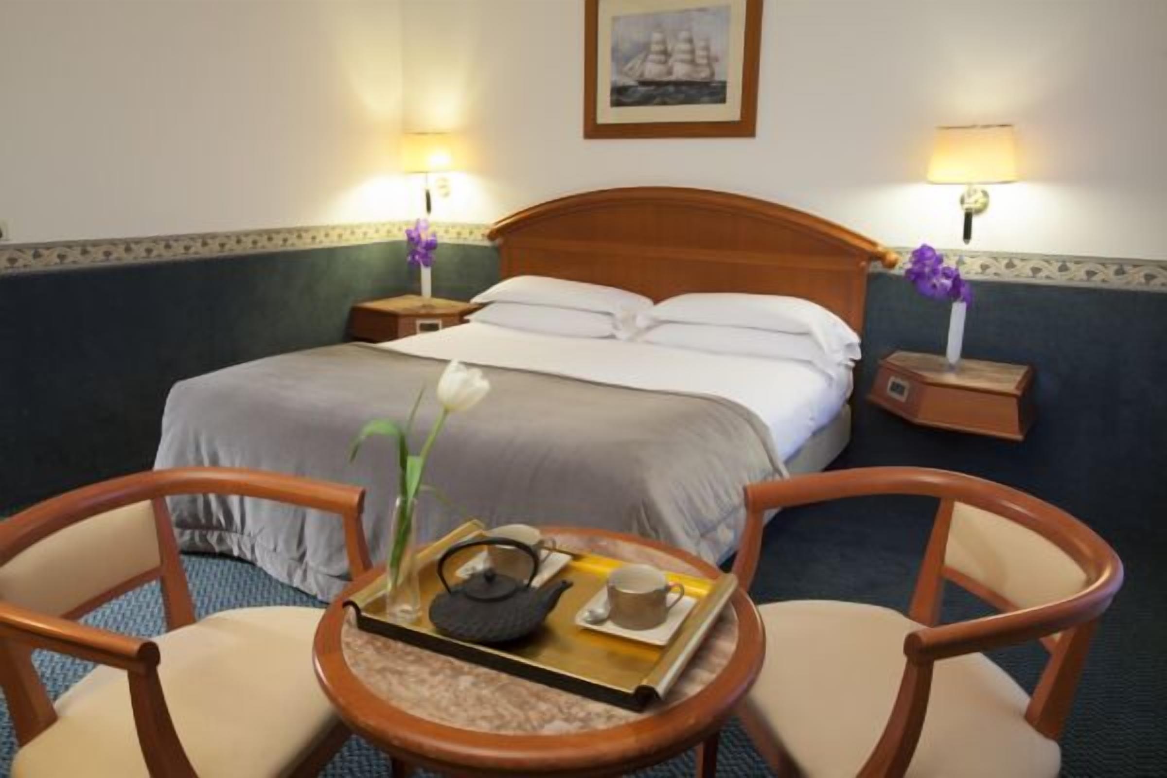 Ein Hotelzimmer mit einem Bett, zwei Nachttischen mit Lampen, einem kleinen runden Tisch mit Tablett und zwei Stühlen, sowie einem gerahmten Bild an der Wand.