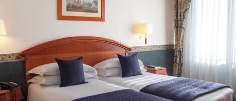 A hotel room with two beds, a wooden headboard, a framed painting on the wall, and a nightstand with a lamp.