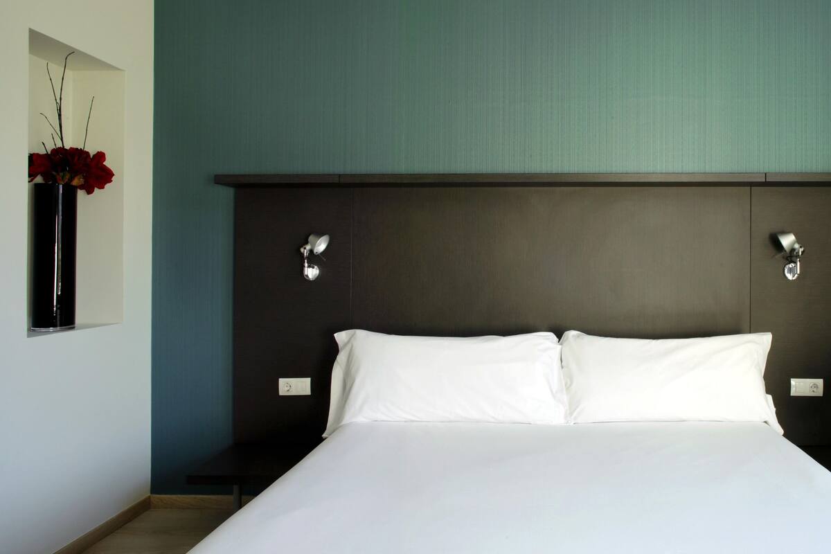 A hotel room with a large bed, a dark headboard, a black chair, and a vase with red flowers on the wall.