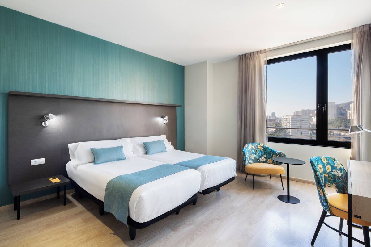 A hotel room with a large bed, two bedside tables, a small round table, a chair, and a large window offering a city view.