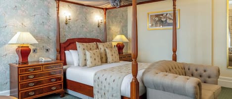 Una cama con dosel de madera, dos mesitas de noche con lámparas, un sillón tapizado en rojo y un papel tapiz estampado con pájaros.