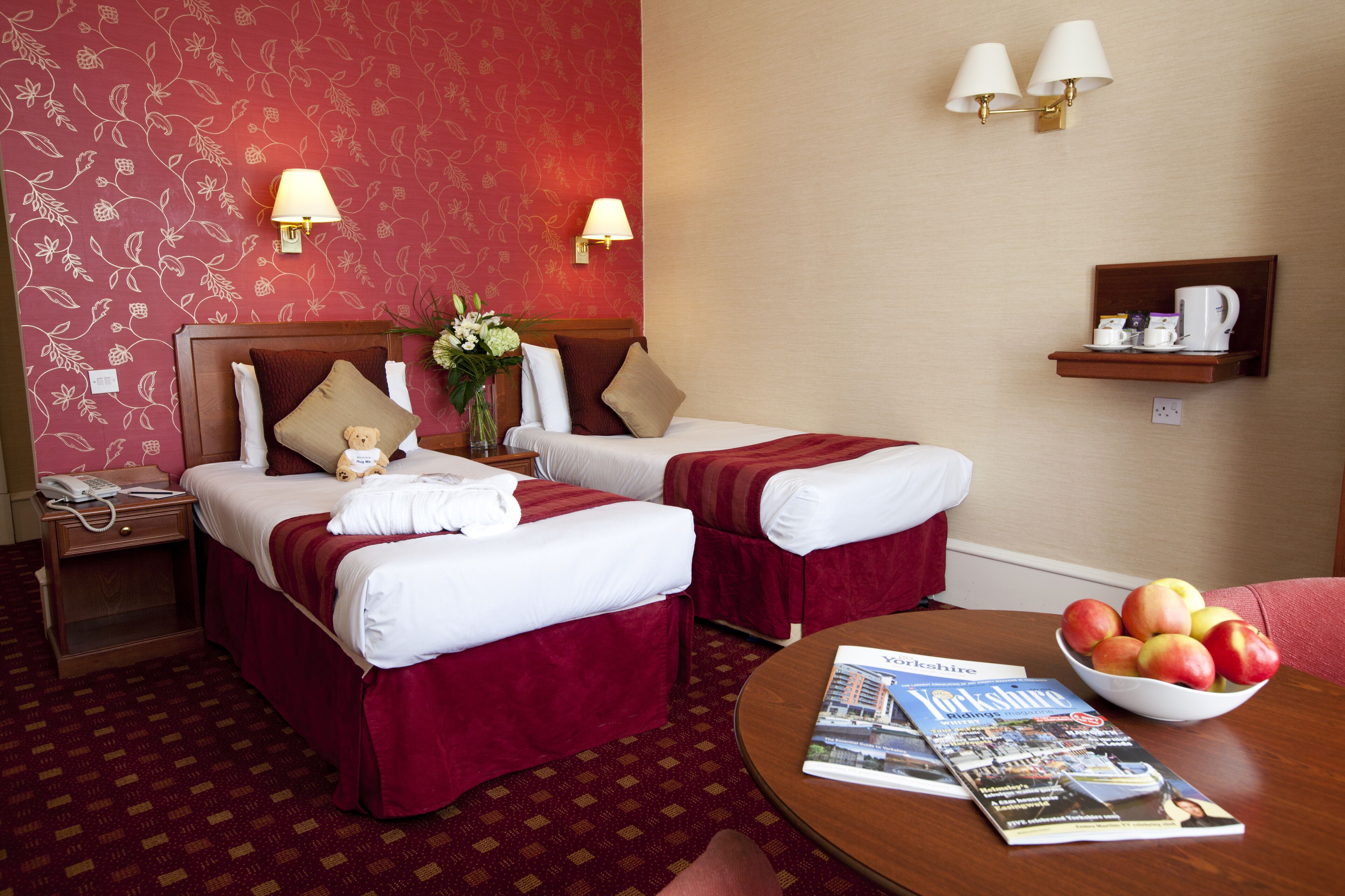 A hotel room with two beds, a nightstand, a table with a bowl of fruit, and a magazine.