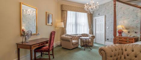 Habitación de hotel con escritorio, dos sillones, sofá, una mesa redonda, una araña y una cama con dosel.