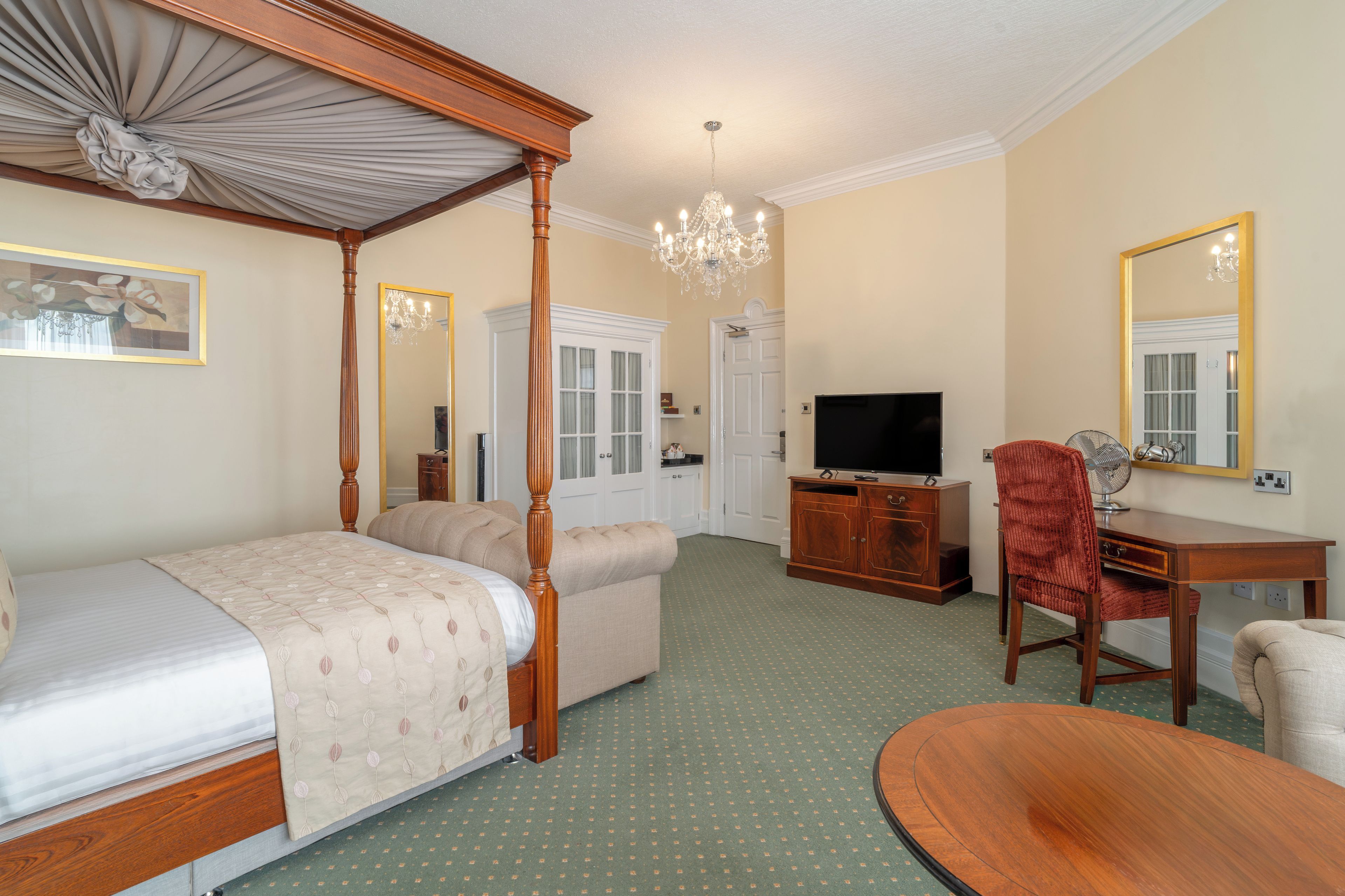 A bedroom with a four-poster bed, a sitting area, a television, a desk, and a mirror.