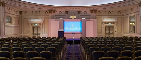 A conference room with rows of chairs, a stage with a screen displaying 'MIDLAND HOTEL', and a carpeted floor.