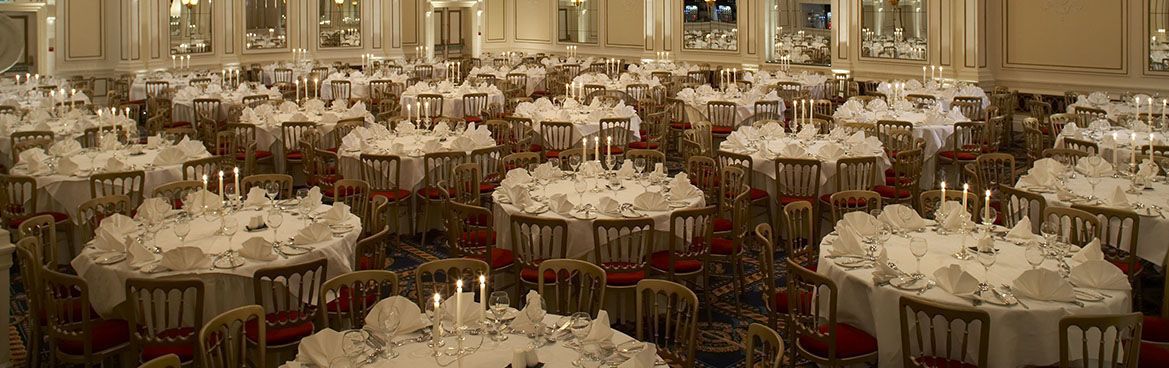 Un gran salón de banquetes con mesas redondas dispuestas para cenar, cada una con mantel blanco y velas.