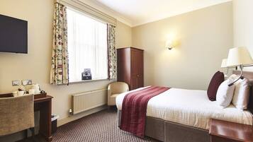 A hotel room with a bed, a chair, a desk, a radiator, a television, and a window with curtains.