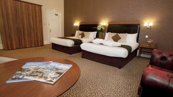A hotel room with two beds, a wooden wardrobe, a round coffee table with magazines, a red leather chair, and a patterned carpet.