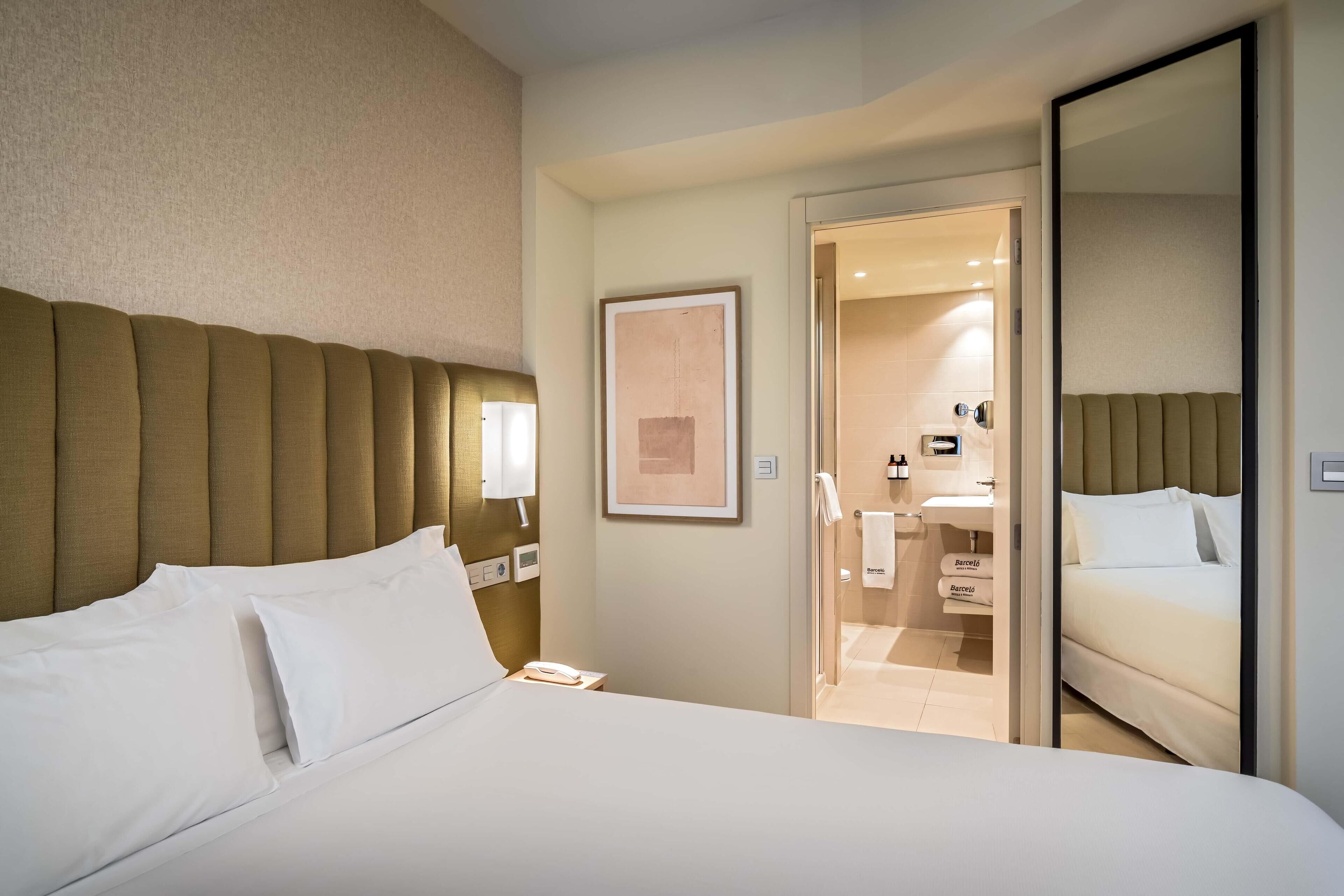 A hotel room with a large bed, a bedside table with a lamp, a framed picture on the wall, and a bathroom visible through an open door.