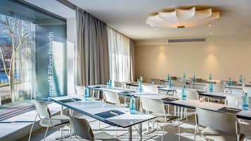 Un moderno salón de conferencias con paredes de vidrio, sillas y mesas blancas, equipado con botellas de agua y vasos.