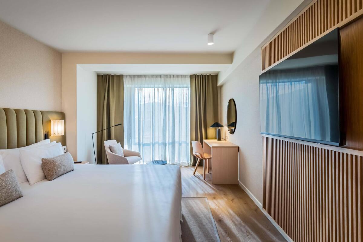 A hotel room with a large bed, a desk, a chair, a TV, and a window with curtains.