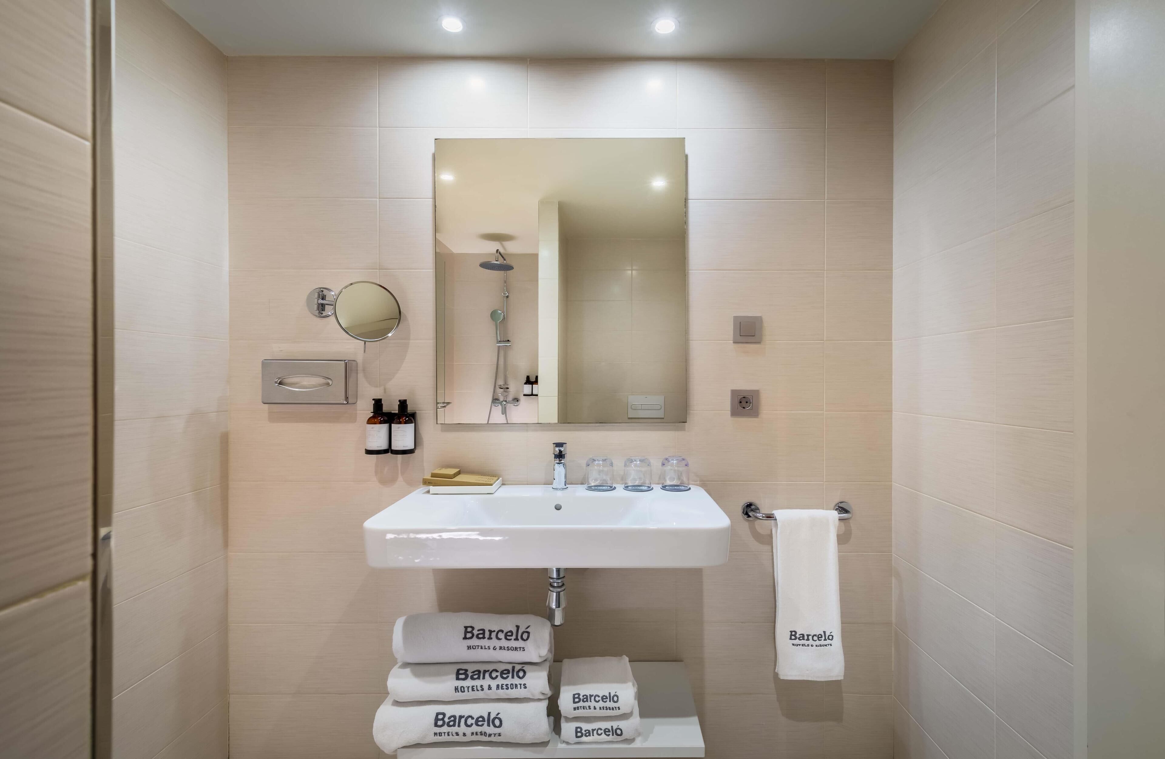 A modern bathroom with a white sink, a mirror, and a shower. There are towels and toiletries on the sink.