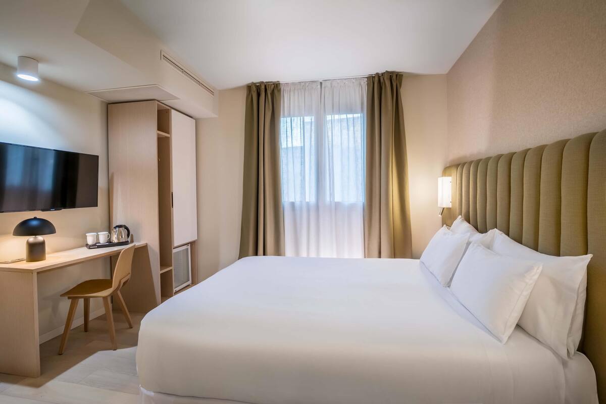 A hotel room with a large bed, a desk with a chair, a television, and a window with curtains.