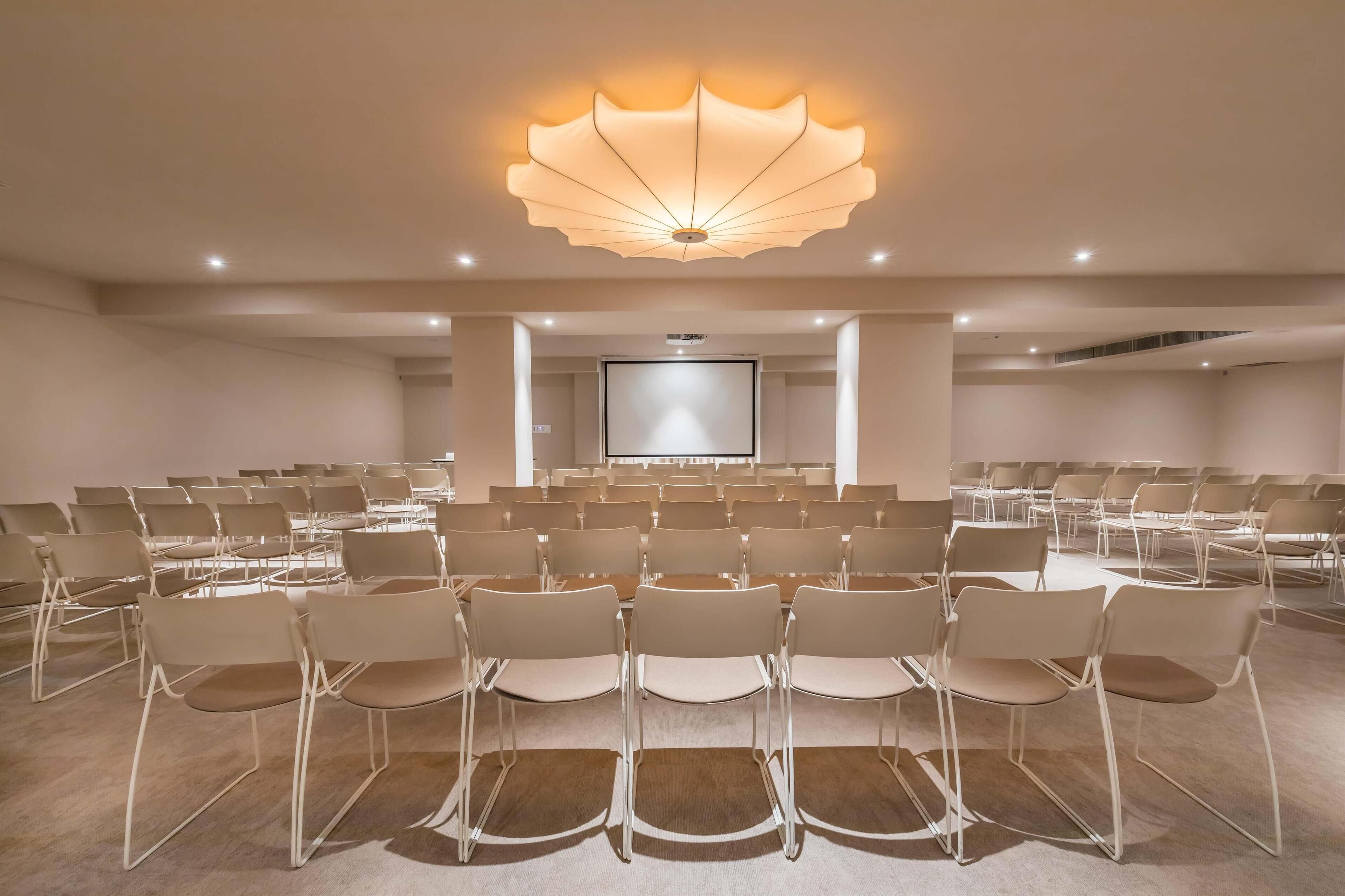 Uma sala de conferências com fileiras de cadeiras, uma tela grande na frente e um lustre de teto único.