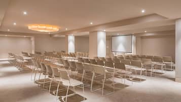 Un salón de conferencias con filas de sillas, una pantalla de proyector e iluminación empotrada en el techo.