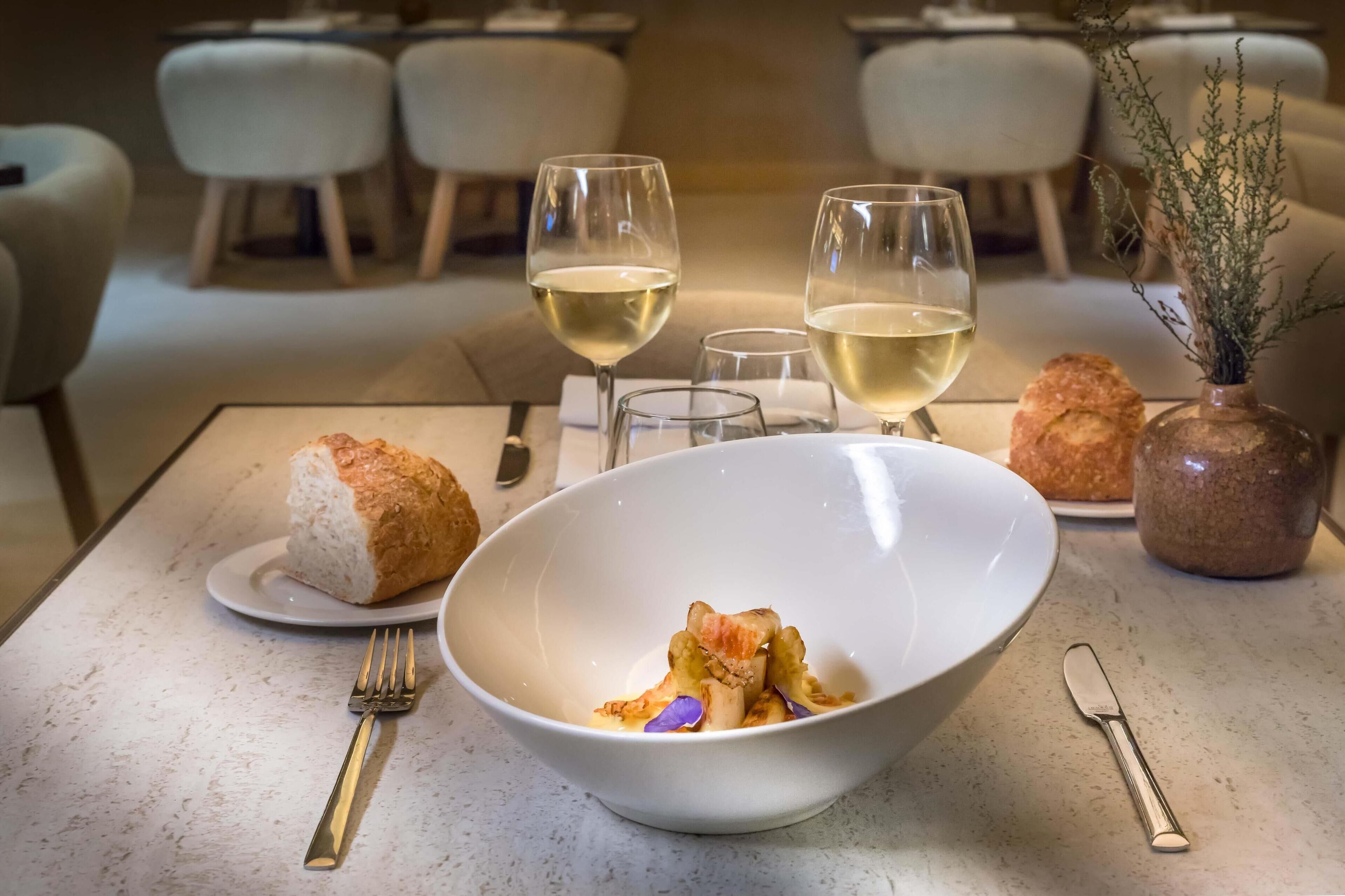 A dining table with a white bowl containing food, two glasses of white wine, a fork, a knife, a loaf of bread, and a small vase with dried plants.