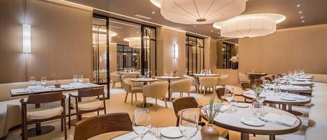 Un comedor amplio con mesas redondas, iluminación colgante moderna y una paleta de colores neutros.