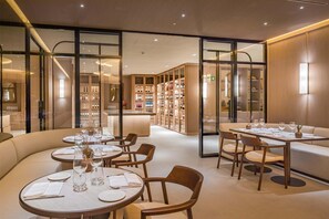 A modern restaurant with round tables and cushioned seating, a wine cellar in the background, and a glass partition separating the dining area from the rest of the space. - Barceló Bilbao Nervión (Bilbao)