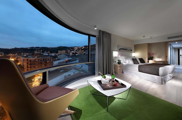 A modern hotel room with a curved balcony, a bed, a sofa, and a coffee table with a bottle and glasses. - Barceló Bilbao Nervión (Bilbao)
