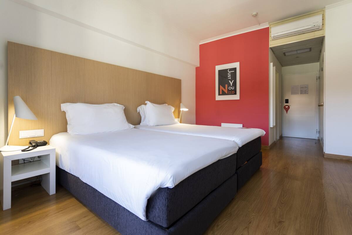 A hotel room with a large bed, bedside tables, a red accent wall, and a door leading to another room.