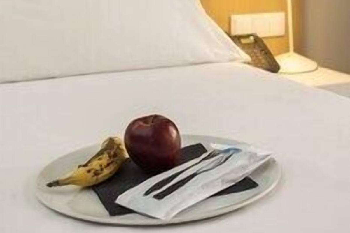 A neatly made bed with a pillow, a bedside lamp, and a plate with a banana and an apple.