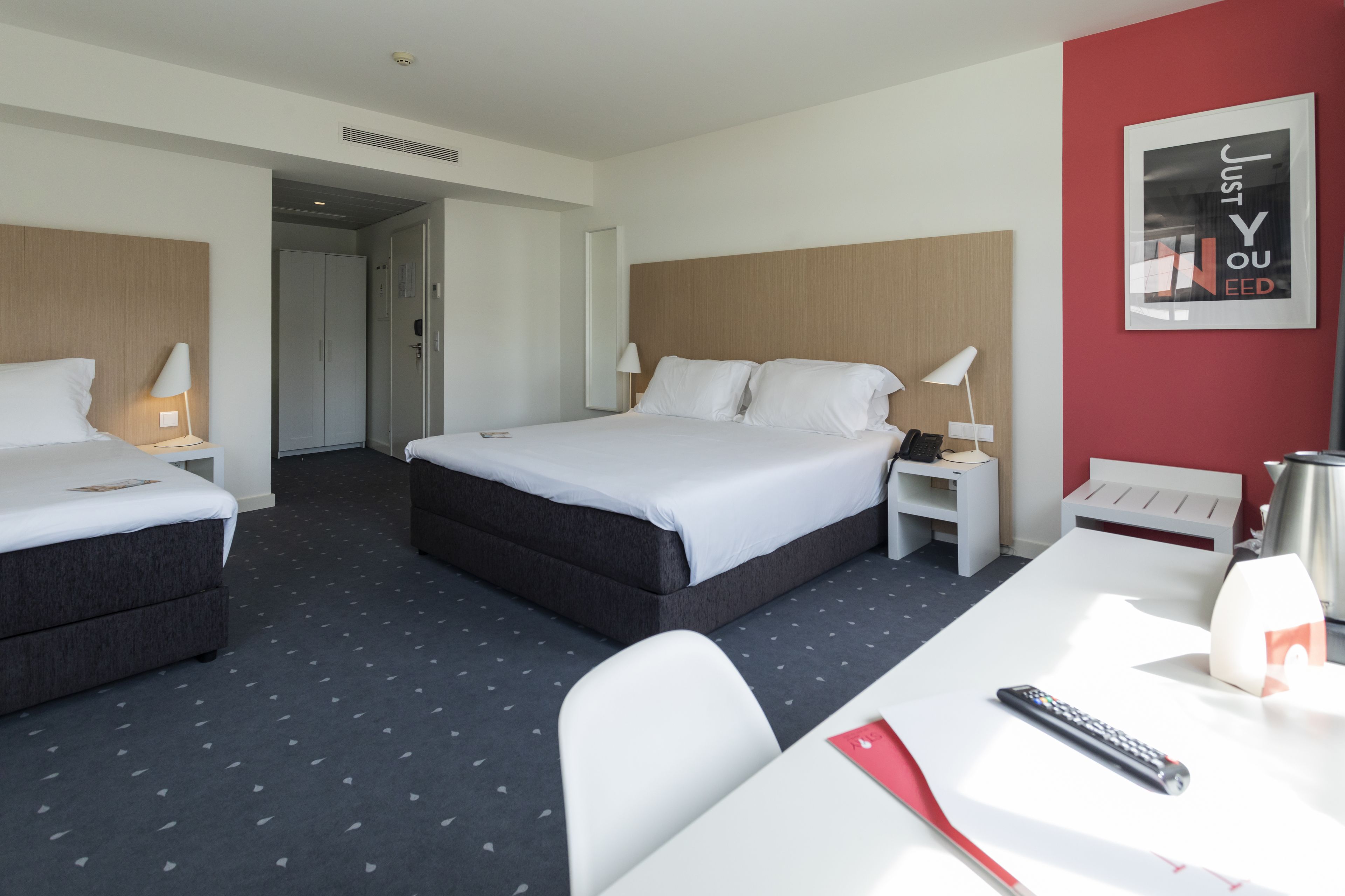 Een hotelkamer met een groot bed, twee nachtkastjes, een bureau en een stoel. Op de muur hangt een lijst en op het bureau ligt een afstandsbediening.