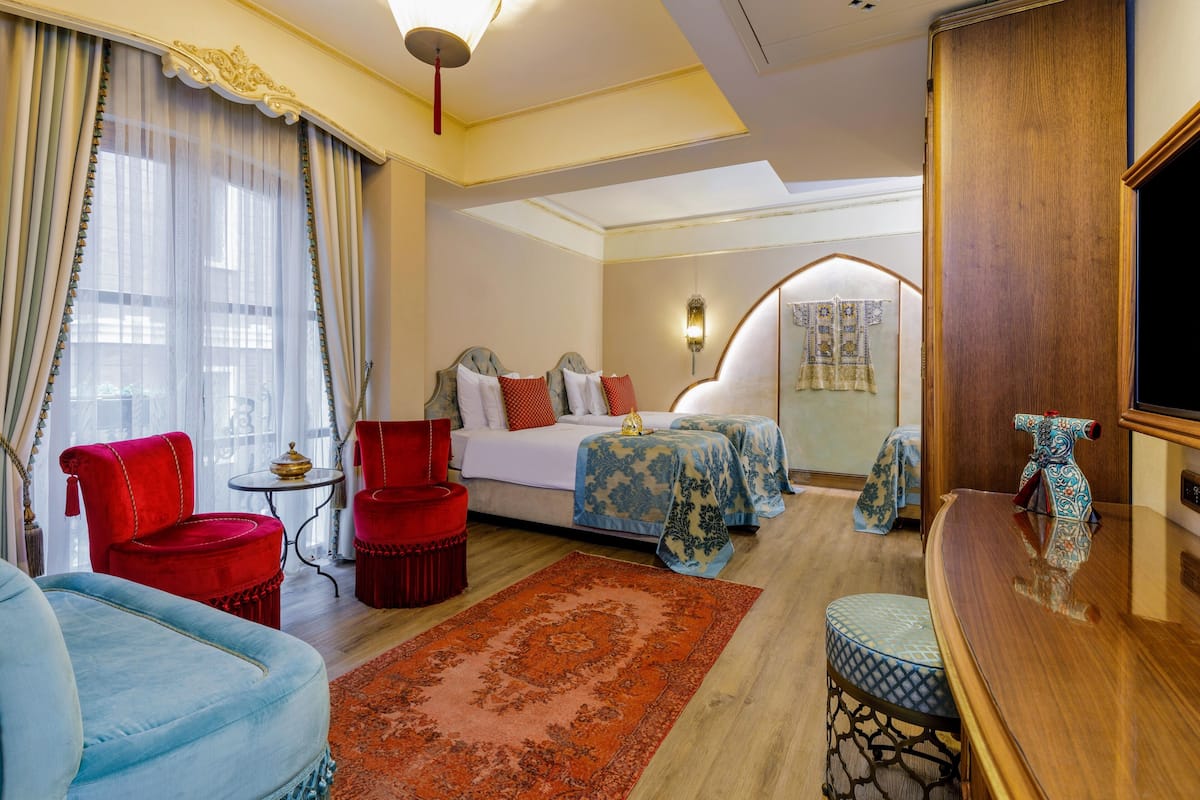 A hotel room with a bed, two red chairs, a small table, a wooden side table, a TV, and a decorative vase.