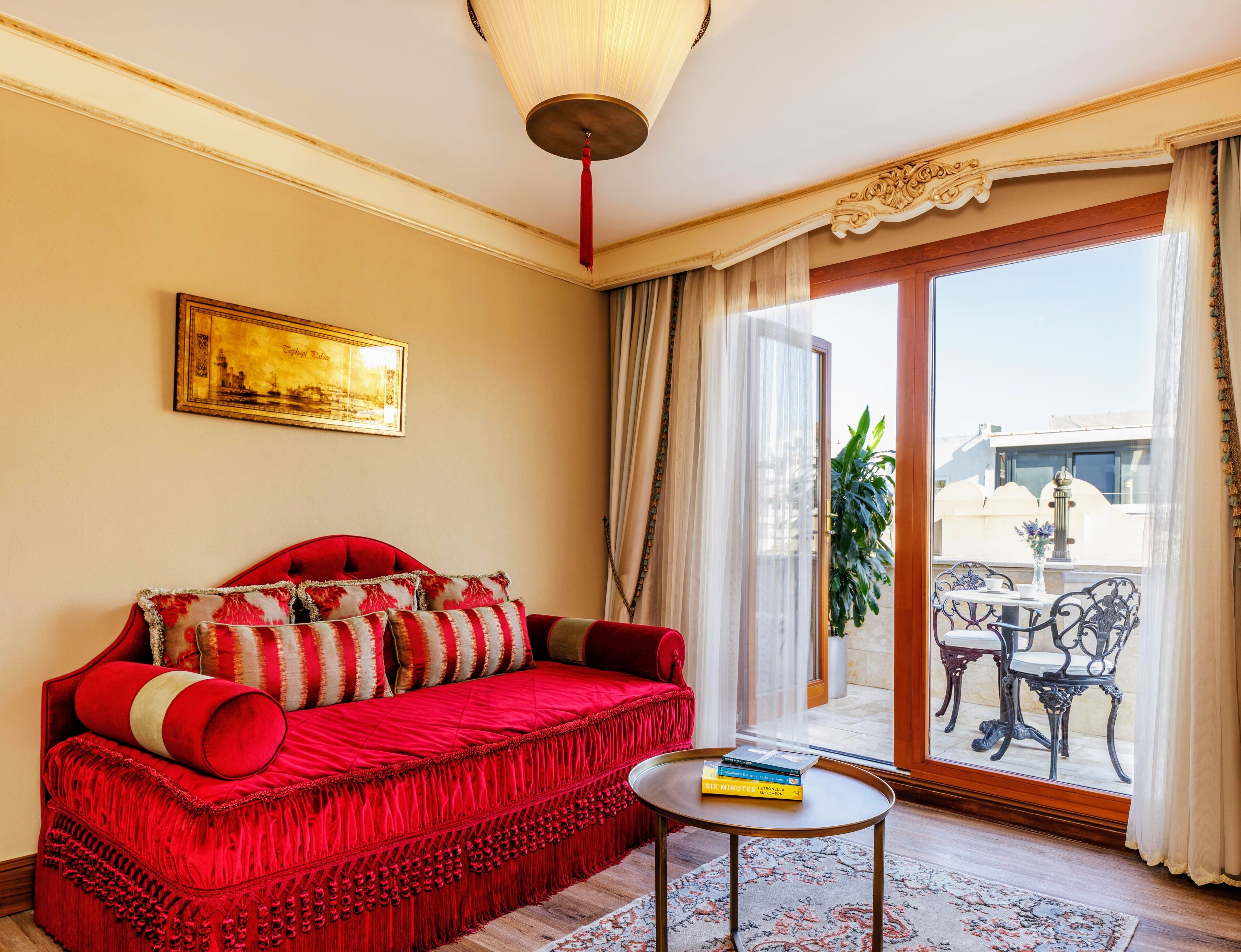 A red sofa with patterned cushions, a small round table with books, a framed picture on the wall, and a view of the outside balcony.