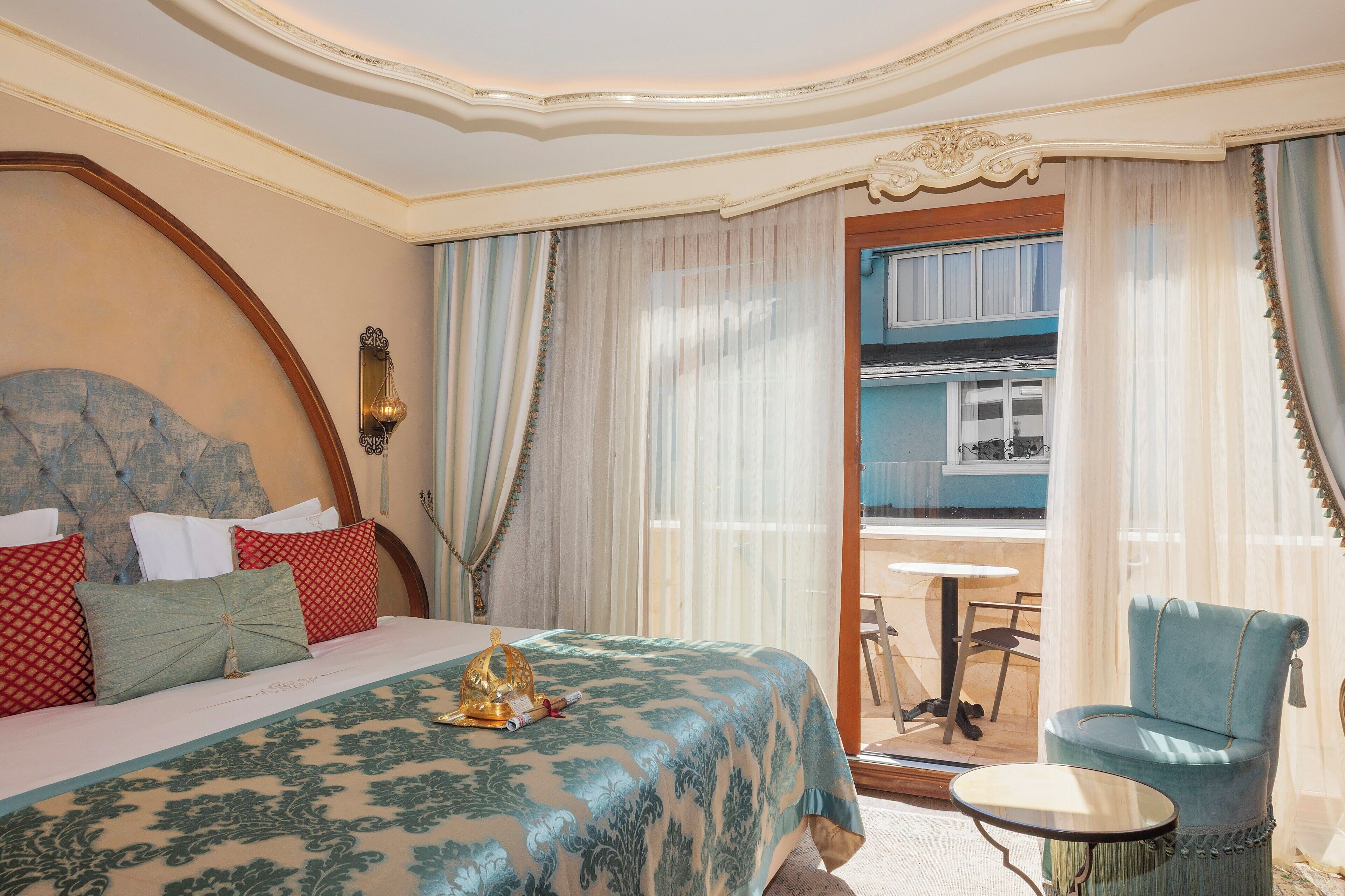 A hotel room with a large bed, a small round table, a blue armchair, and a balcony with a view.