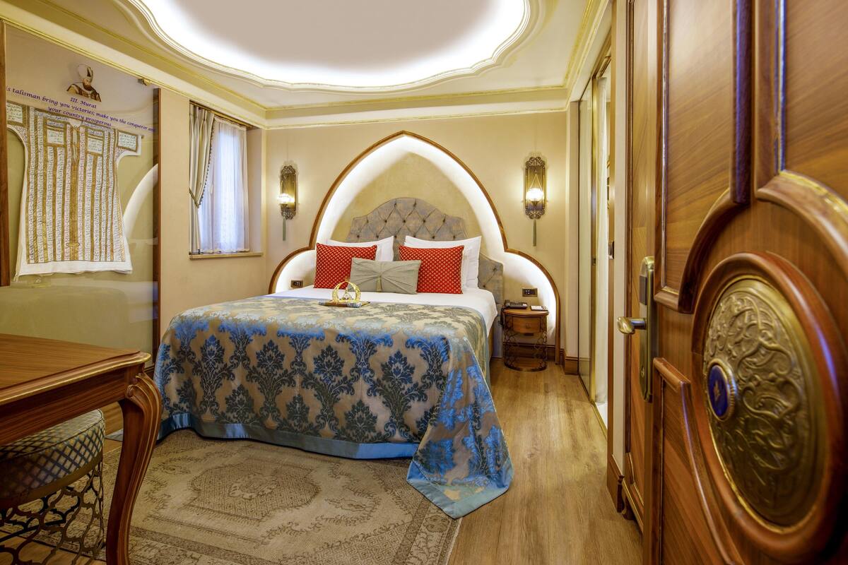 A bedroom with a large bed, a wooden nightstand, a wall-mounted lamp, and a decorative mirror.
