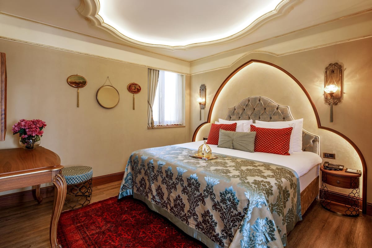 A bedroom with a large bed, a wooden nightstand, a red carpet, and a wooden desk with a vase of flowers.