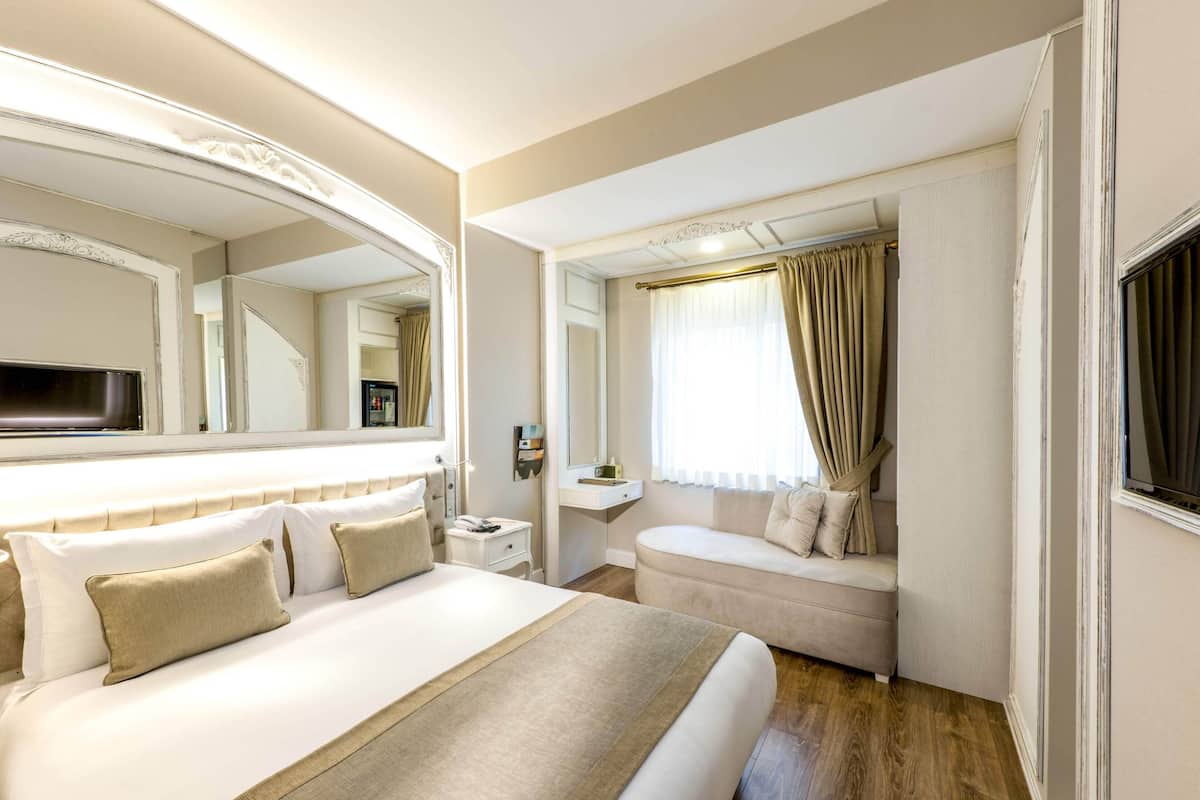 A hotel room with a large bed, a sofa, a bedside table, a mirror, and a window with curtains.