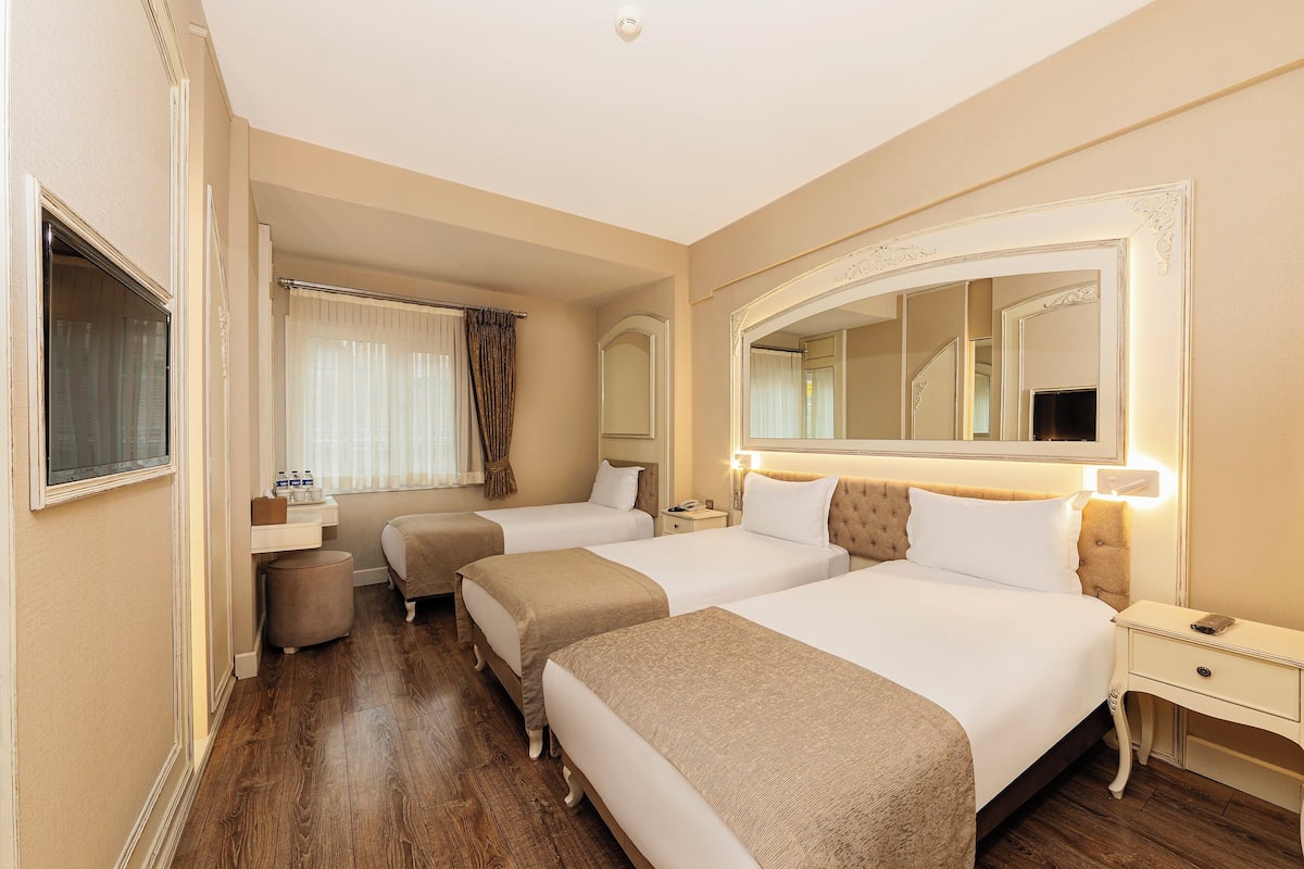 A hotel room with two beds, a large mirror, a bedside table with a lamp, and a window with curtains.