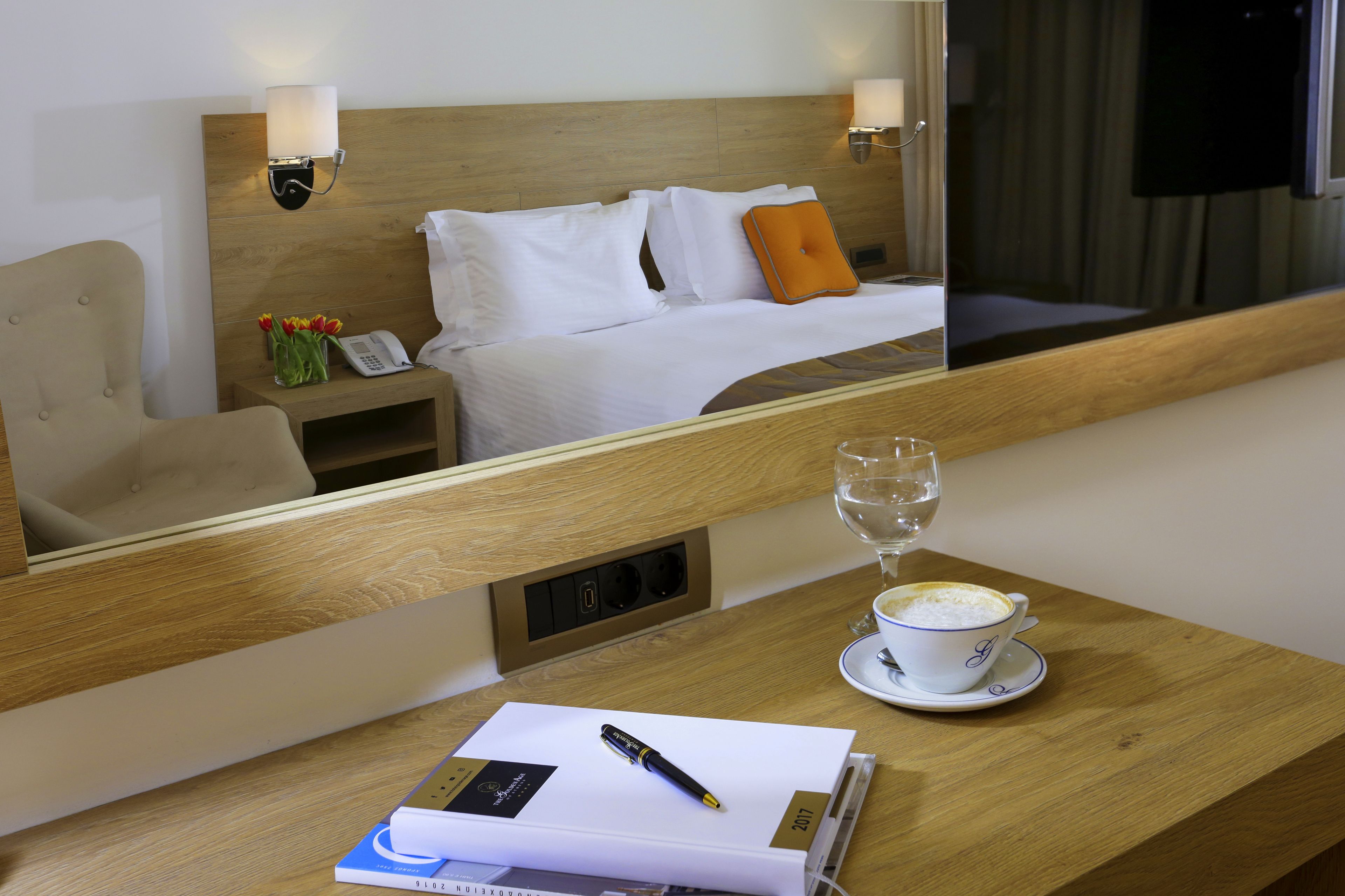 A hotel room with a wooden headboard, a bedside table with a lamp, a cup of coffee, a notebook, and a pen.