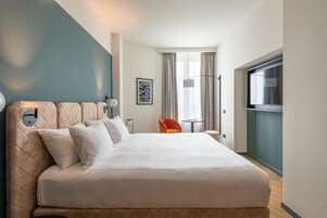 Una camera d'albergo con un letto grande, un tavolino, una sedia, una finestra con tende e una TV.