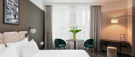 Een hotelkamer met een groot bed, twee groene stoelen, een klein rond tafeltje met een bloemenvaas, een bureau met een lamp, en een lijst met een afbeelding aan de muur.