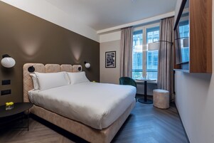 Een moderne hotelkamer met een groot bed, een klein tafeltje, een stoel, een raam met gordijnen en een aan de muur bevestigde televisie.