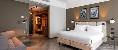 A modern hotel room with a large bed, a seating area, and a walk-in closet.