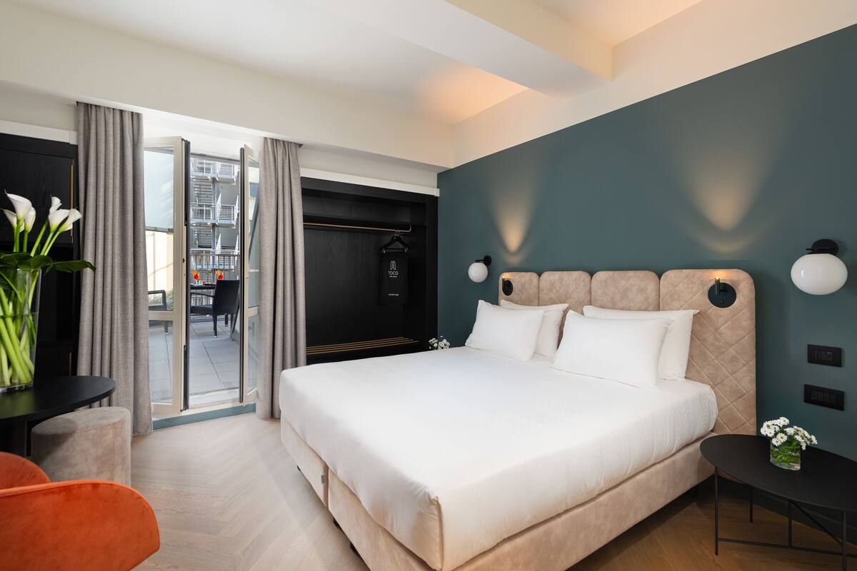 A modern hotel room with a large bed, a dining area with a table and chairs, and a view through a glass door to an outdoor space.