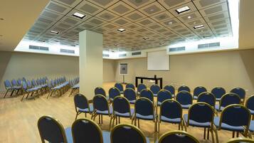 A conference room with rows of blue chairs, a projector screen, and a ceiling with ventilation panels.