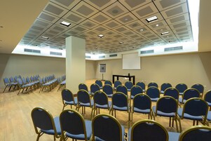 A conference room with rows of blue chairs, a projector screen, and a ceiling with ventilation panels.