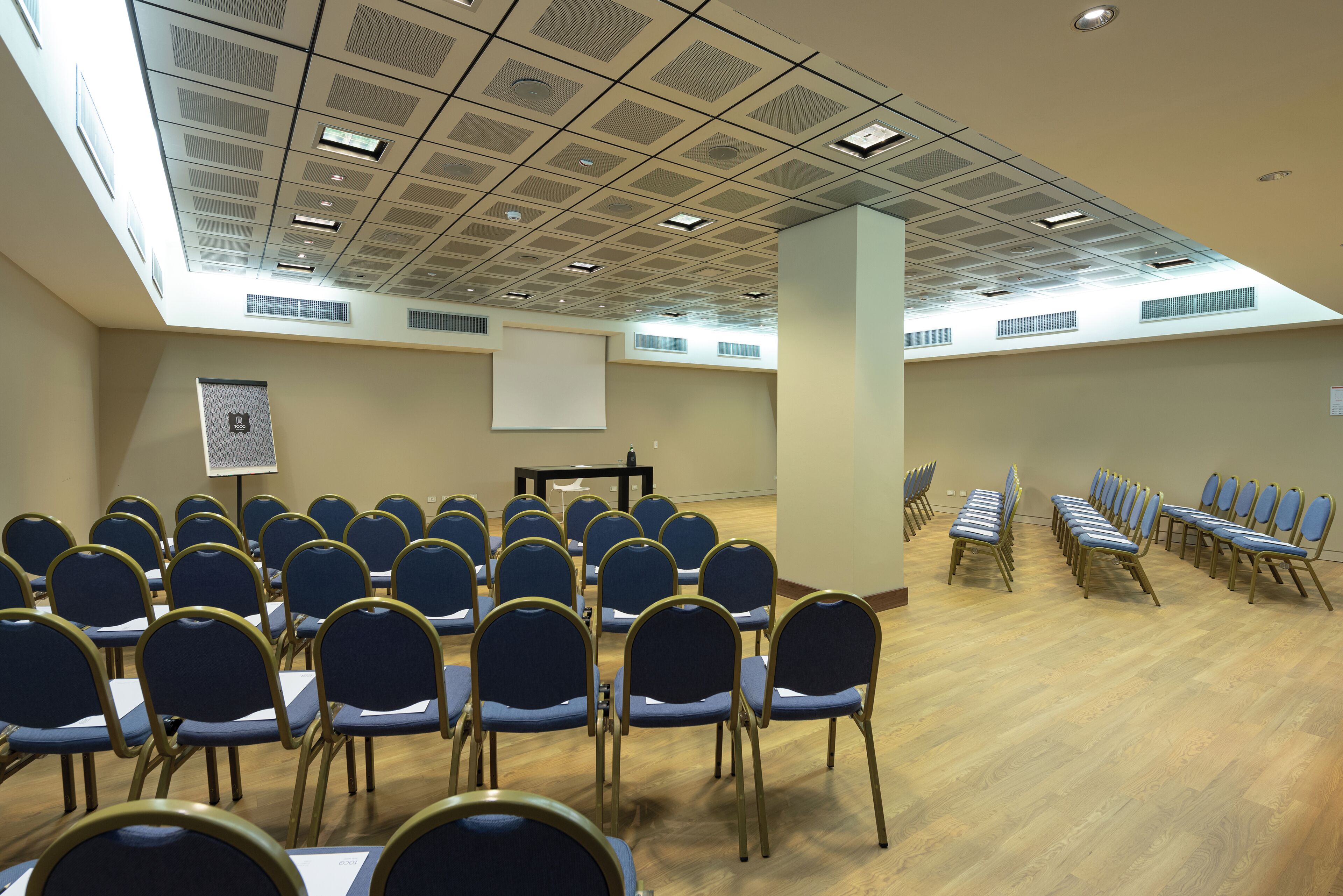 A conference room with rows of chairs, a projector screen, and a ceiling with recessed lighting.