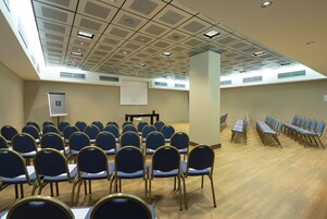 Een vergaderzaal met rijen stoelen, een scherm voor een beamer en een plafond met inbouwspots.